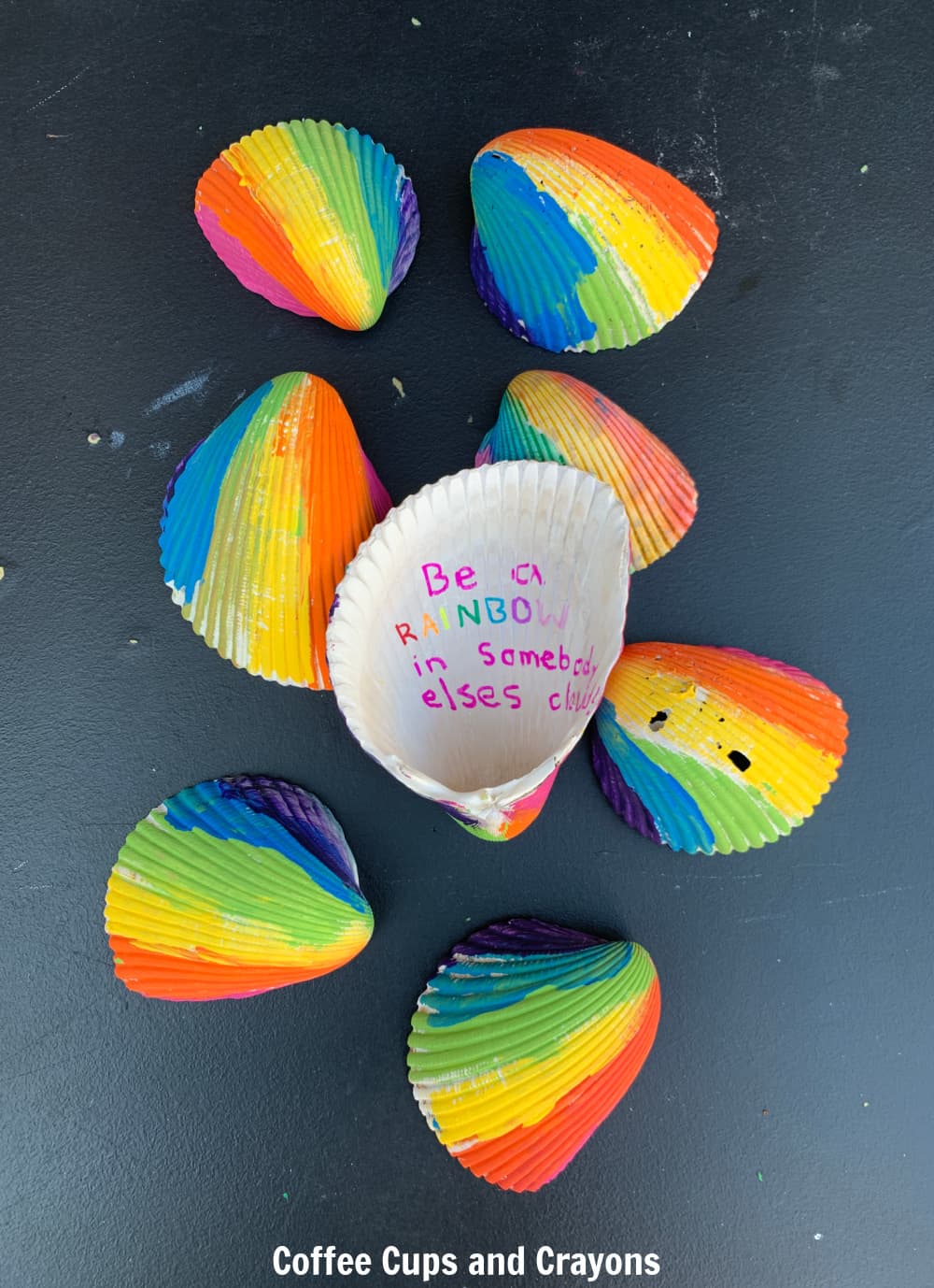 rainbow painted seashells on a black background with one turned over on top and the words "Be a rainbow in someone's else cloud" written on it